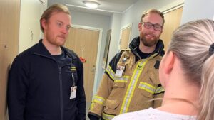 Två brandmän i en korridor. Kvinna med ryggen vänd mot kameran.