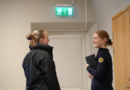 Två kvinnor står och ler mot varandra, den ena är iklädd uniform. I bakgrunden syns utrymningsskylt.