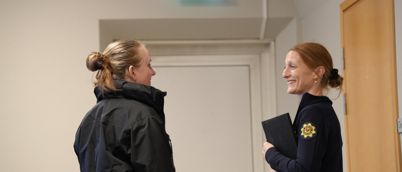Två kvinnor står och ler mot varandra, den ena är iklädd uniform. I bakgrunden syns utrymningsskylt.
