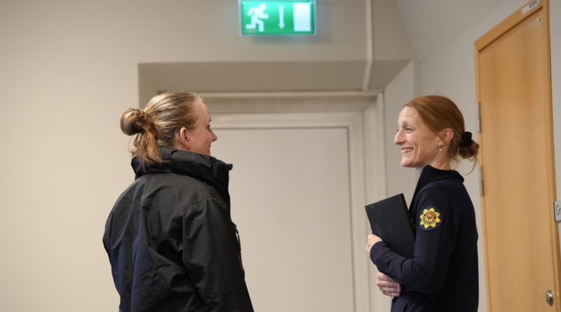 Två kvinnor står och ler mot varandra, den ena är iklädd uniform. I bakgrunden syns utrymningsskylt.